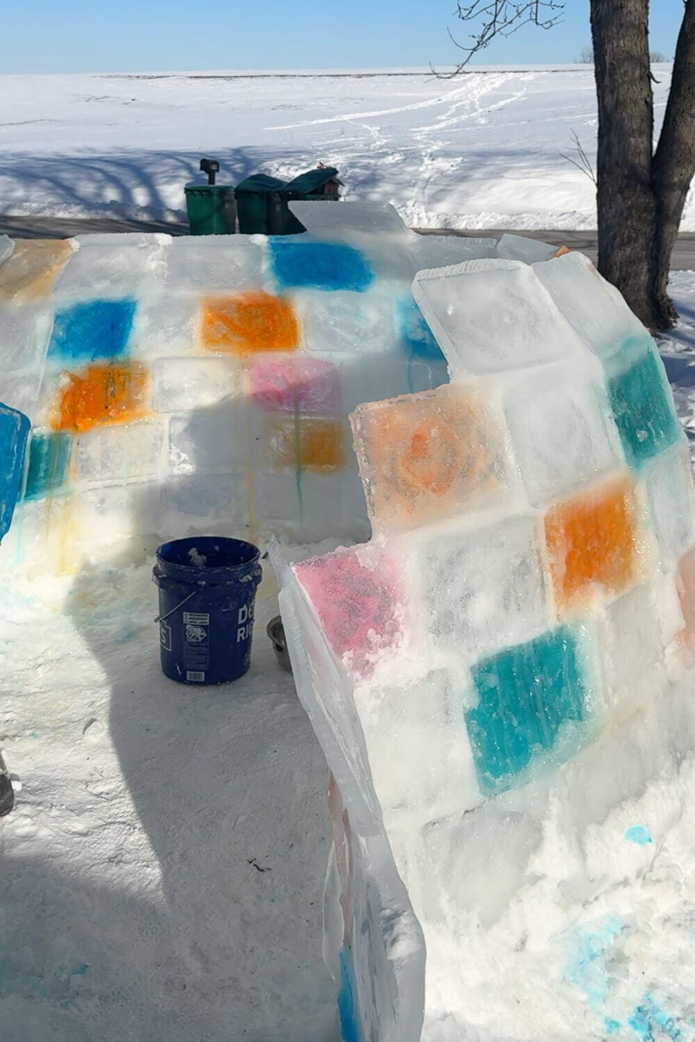 Daytime view of a backyard igloo in progress made from clear and colored ice blocks, showing the entrance opening and stacked walls before finishing the dome for a how to build an igloo tutorial