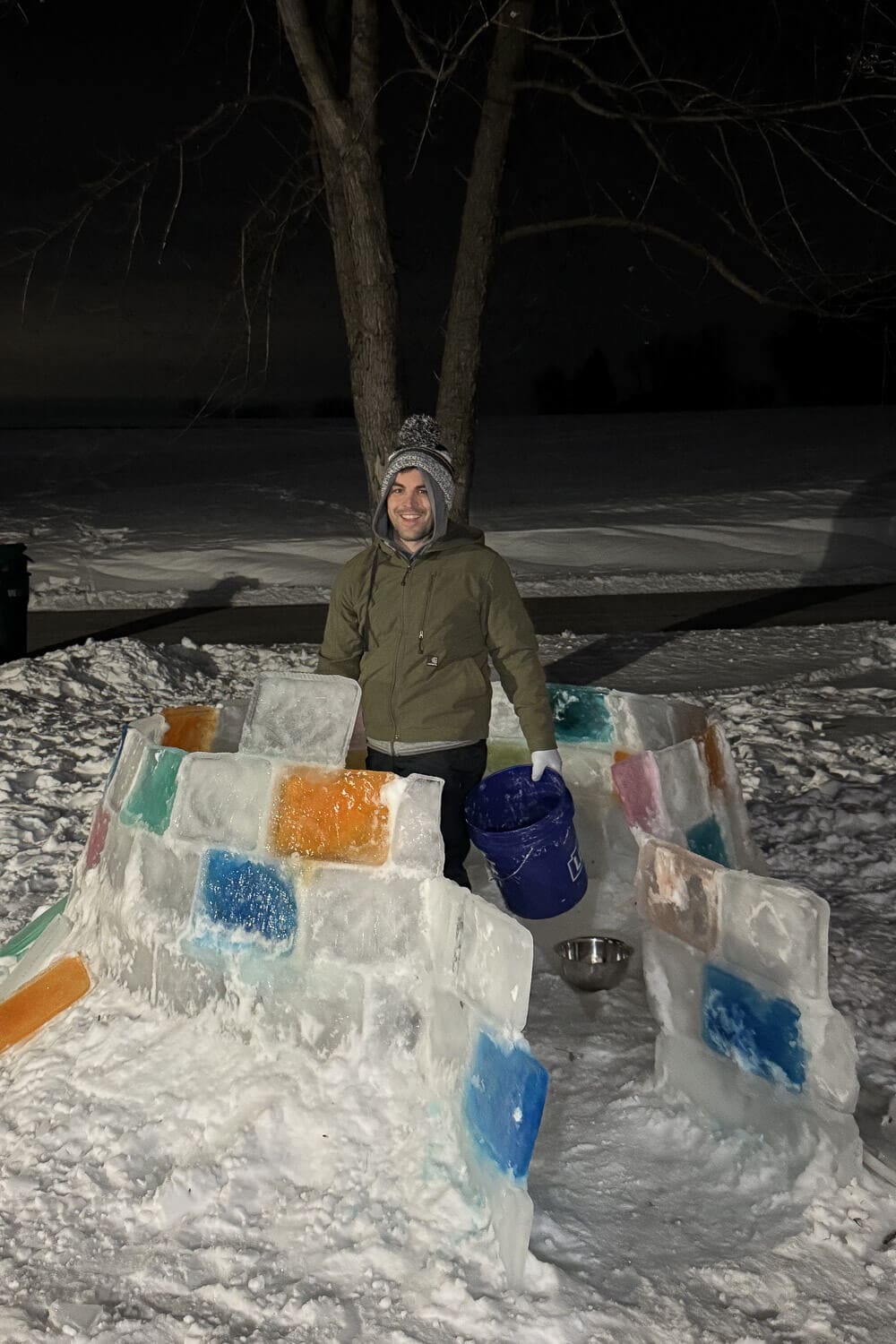 Backyard igloo in progress at night with stacked ice blocks and colorful frozen panels, showing the walls built up before the dome is finished in a how to build an igloo tutorial