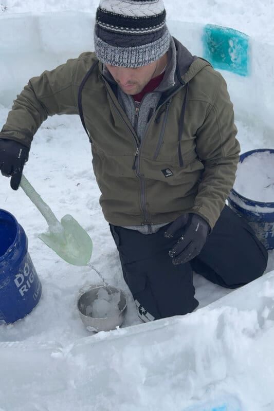 Using a small shovel to stir snow and water into slush for sealing ice blocks, showing how to make an awesome snow fort with homemade slush mortar and ice blocks