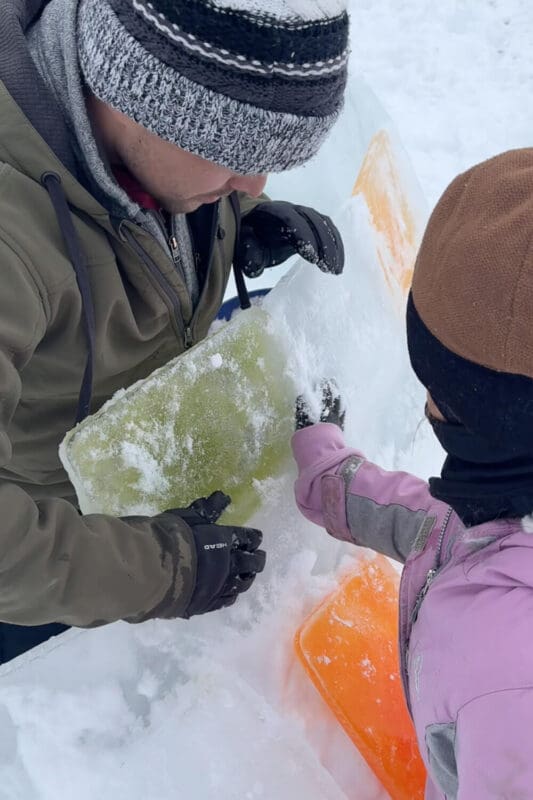 Placing a colored ice block onto an angled igloo row, using slush mortar to bond the blocks as the dome begins to form in a how to build an igloo tutorial