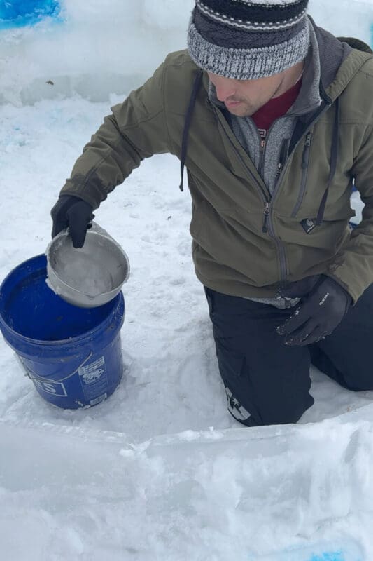Mixing snow and water in a bucket to make slush mortar for a snow fort, creating the perfect consistency to hold ice blocks together.