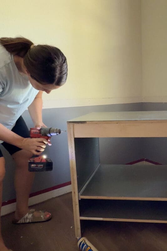 Attaching a rear support stretcher to the back of a DIY base cabinet using a drill before securing it to the wall.