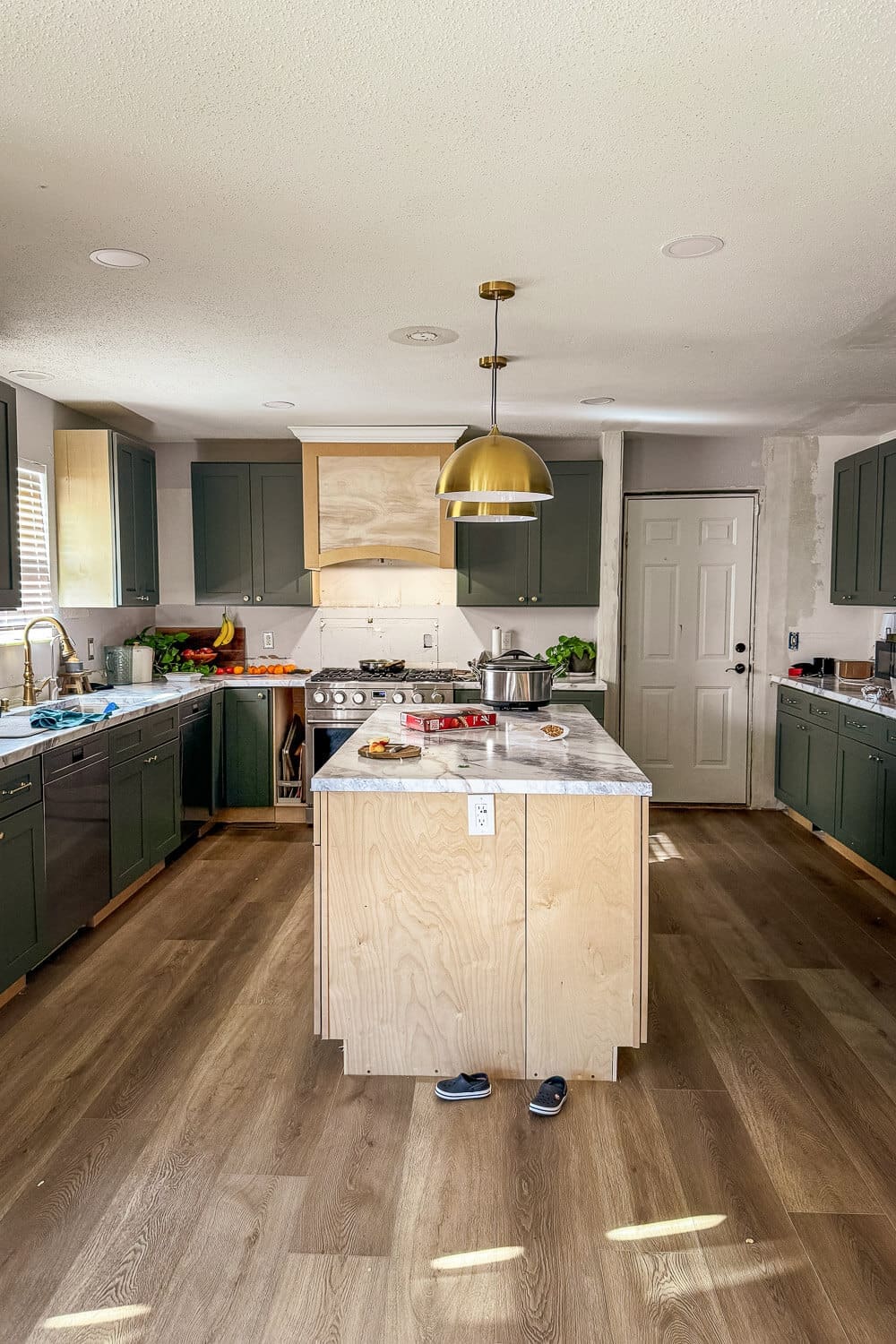 Wide view of my DIY kitchen remodel in progress with dark green cabinets, a plywood island base, marble-look countertops, brass pendant lights, and the range hood and backsplash area still unfinished.