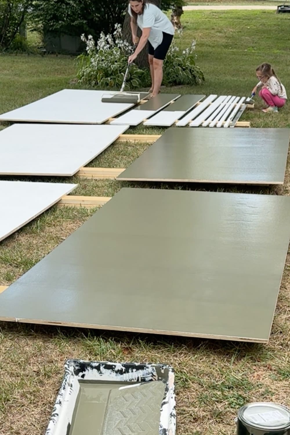 Rolling green paint onto plywood panels laid out in the yard while painting DIY bookcases for our home library, with a helper painting trim pieces nearby.