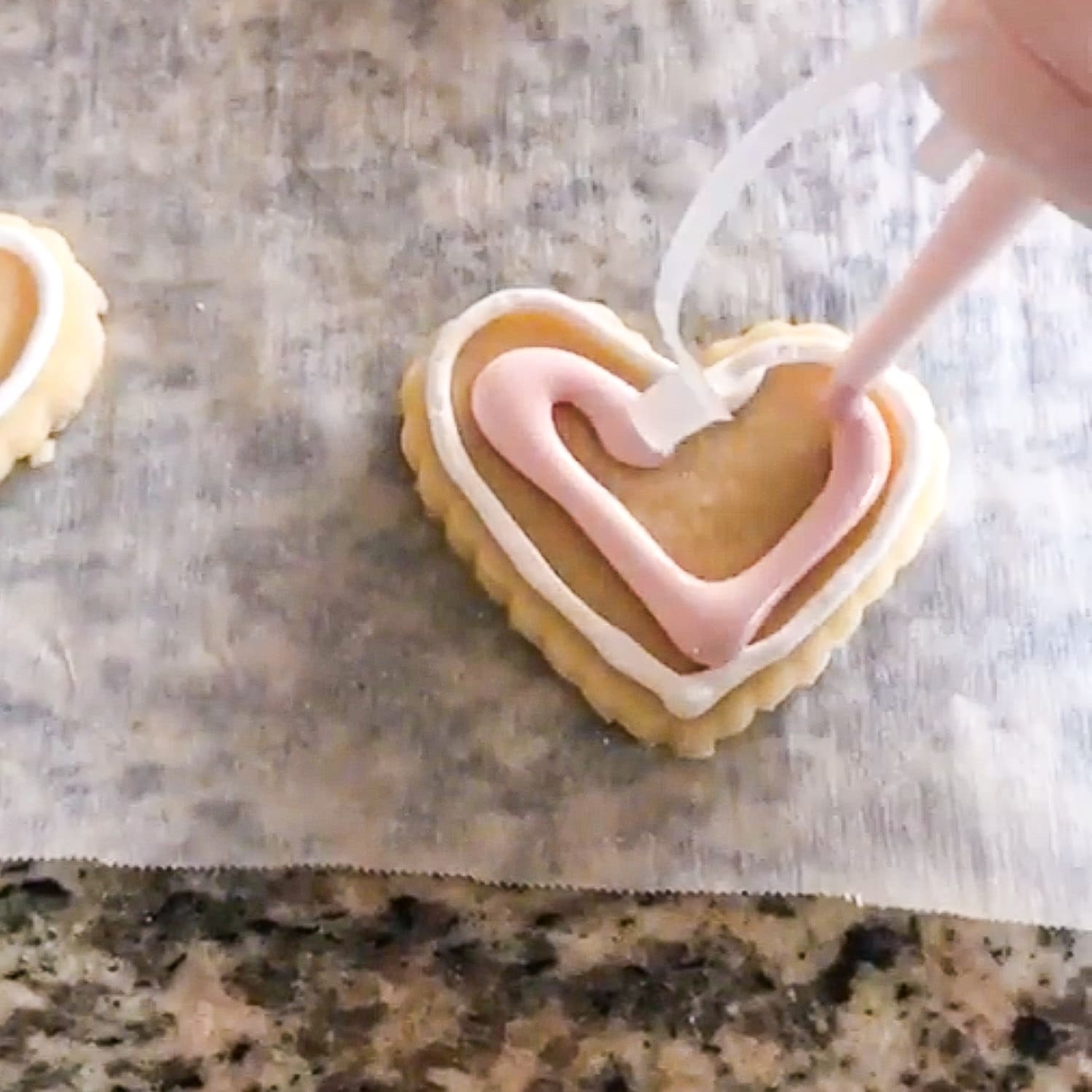 Piping light pink royal icing onto a heart-shaped sugar cookie inside a white outlined border to start flooding the center on parchment paper.