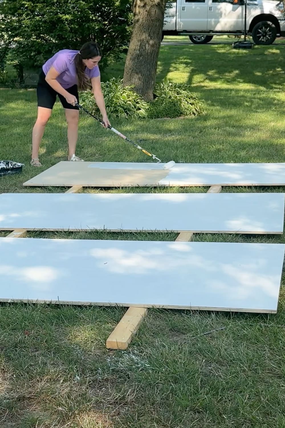 Rolling primer onto long plywood boards laid out in the yard, prepping the panels for painted DIY bookcases for our cozy home library.