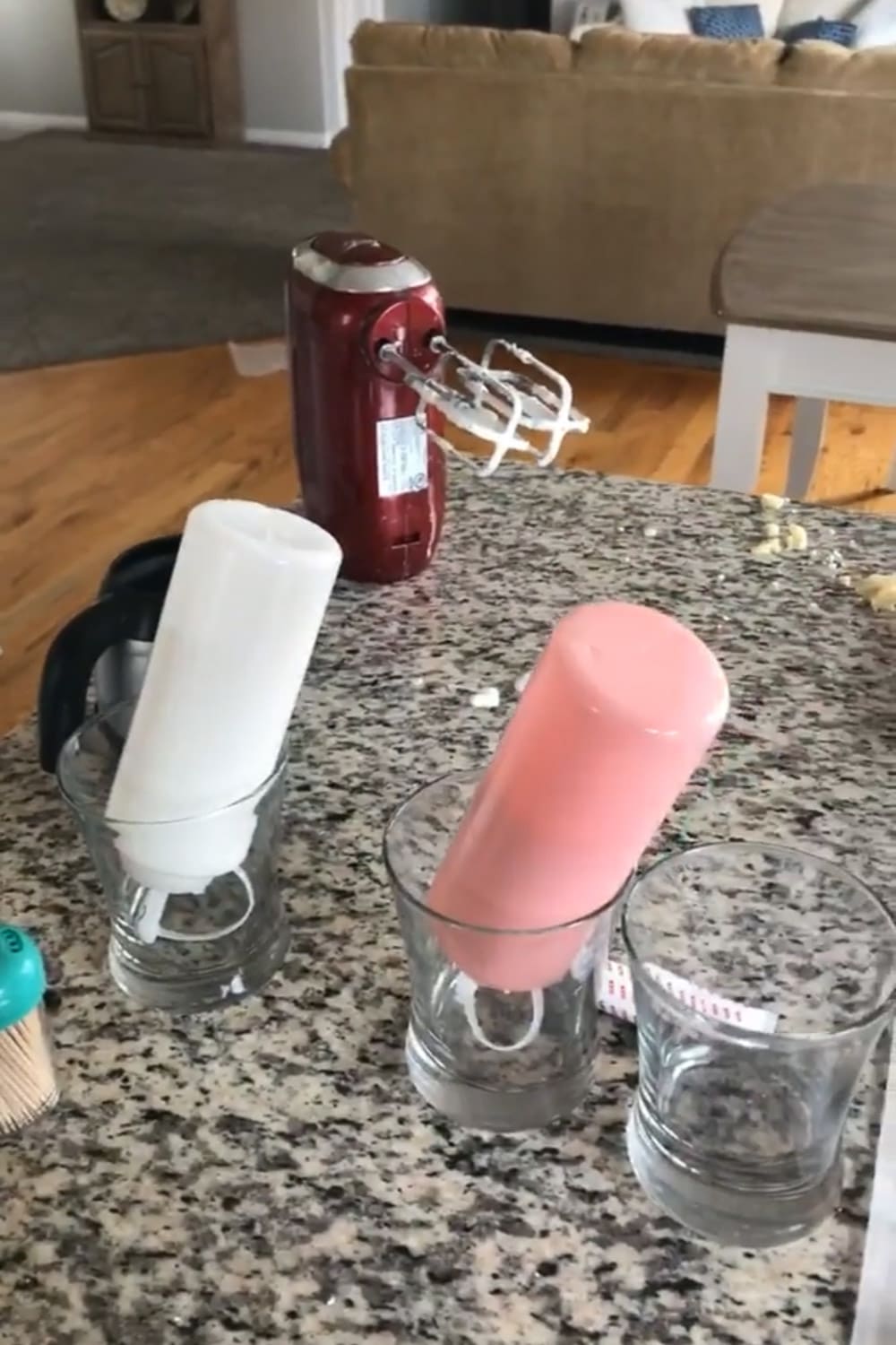 Pink and white squeeze bottles of royal icing drying upside down in drinking glasses on a granite countertop, with a hand mixer in the background.