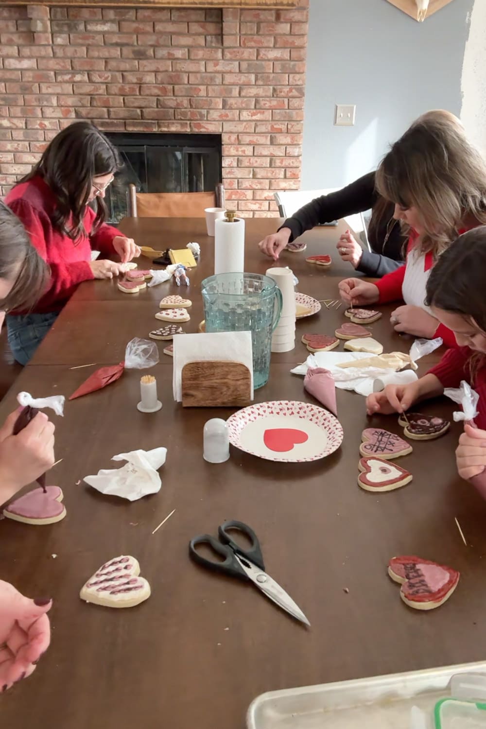 Having a decorating Valentine cookies party at a dining table using piping bags with finished pink, red, and white designs spread out to dry.