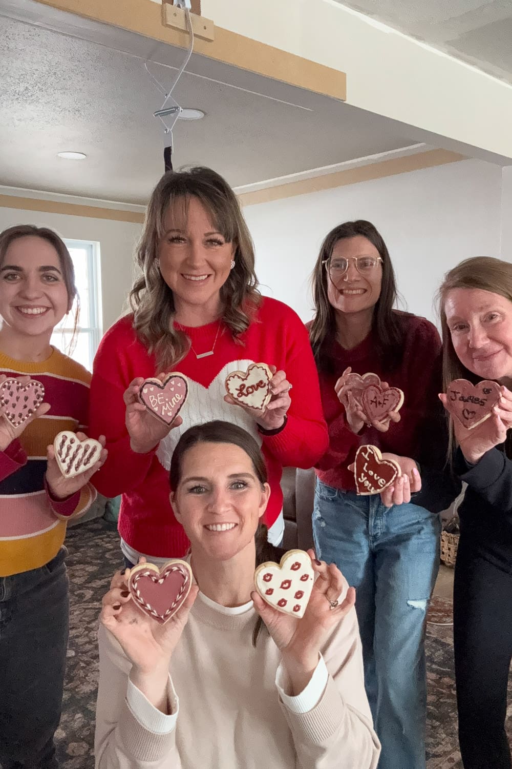 Group of women holding Valentine cookies decorated, showing finished designs like “Be Mine” and “I love you.”