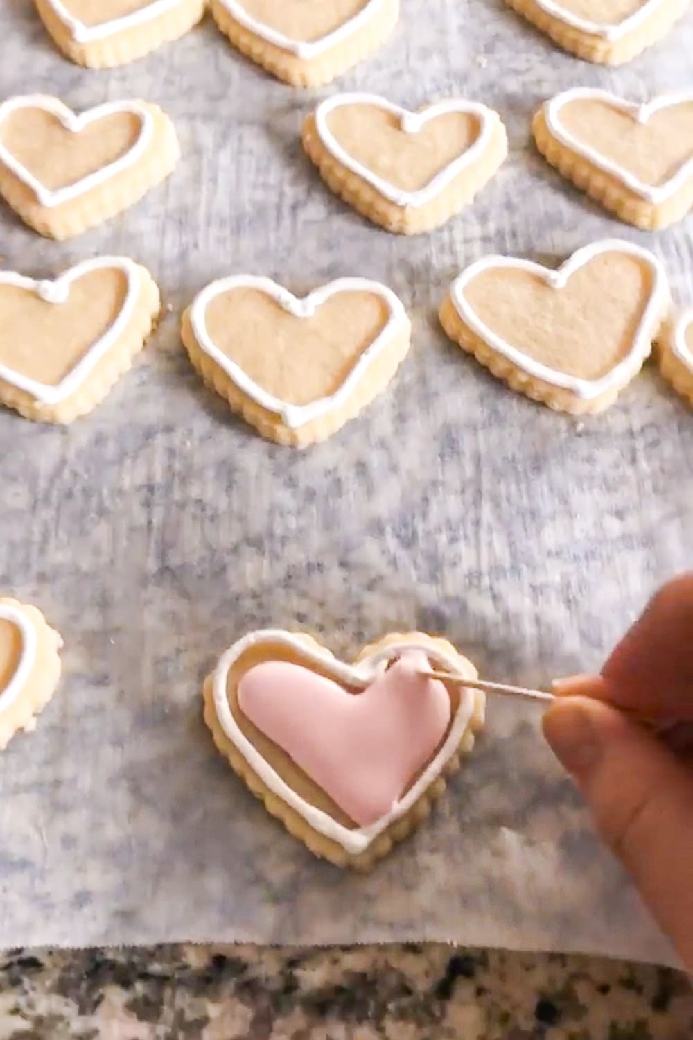 Using a toothpick to smooth and pop bubbles in light pink royal icing on a heart-shaped cookie with a white piped outline, with more heart cookies in the background.
