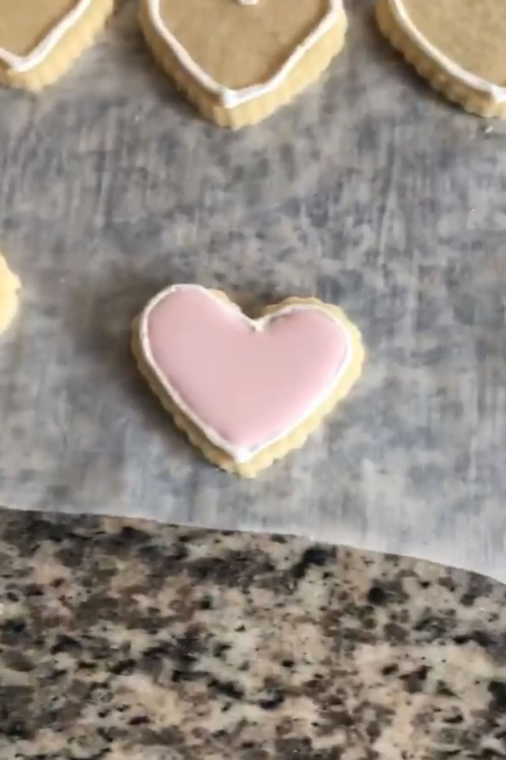 Heart-shaped sugar cookie fully flooded with smooth light pink royal icing on parchment paper, set on a granite countertop.