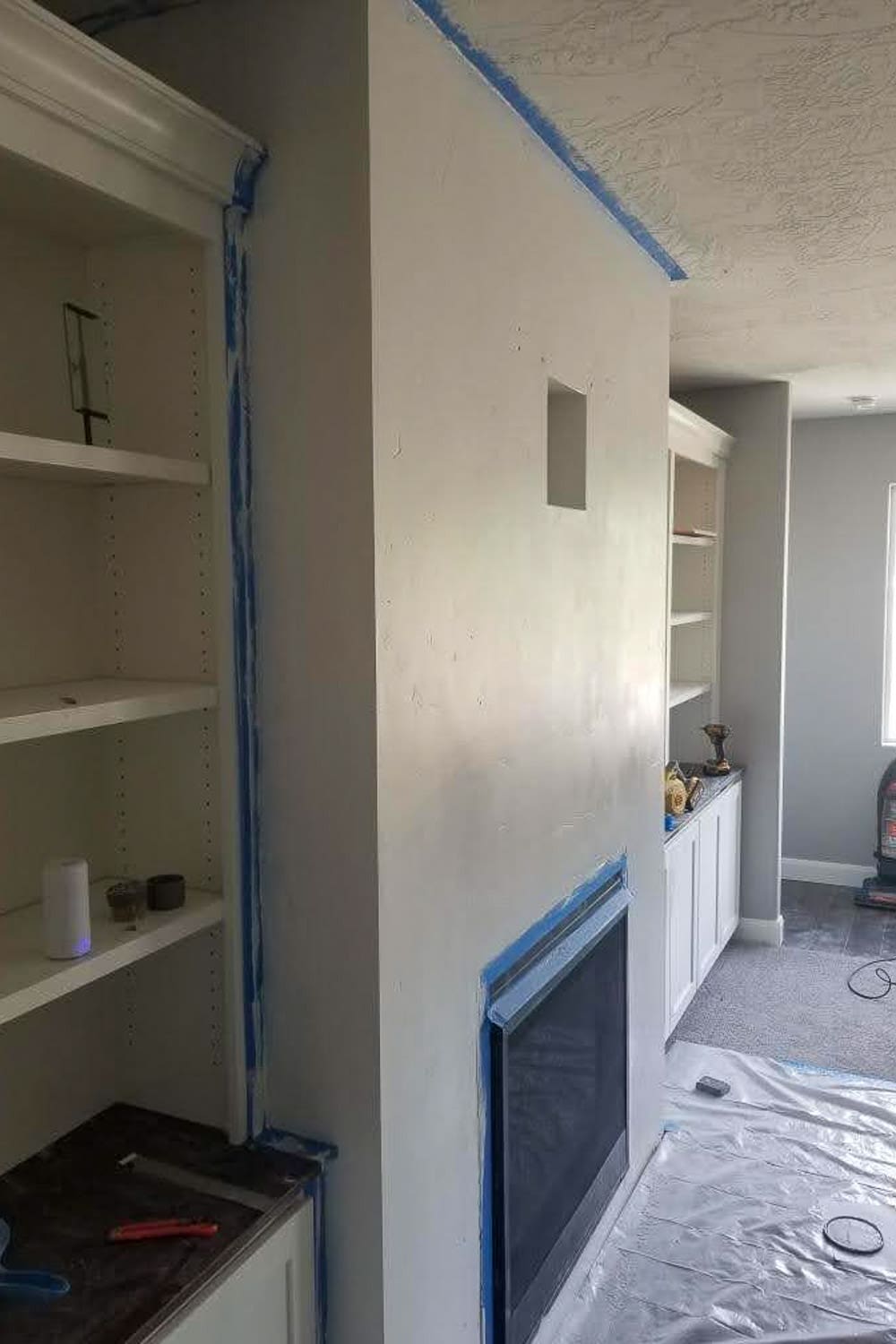 Side angle of primed fireplace wall taped off with blue tape, plastic on floor, recessed media box cutout visible, shop vac in background