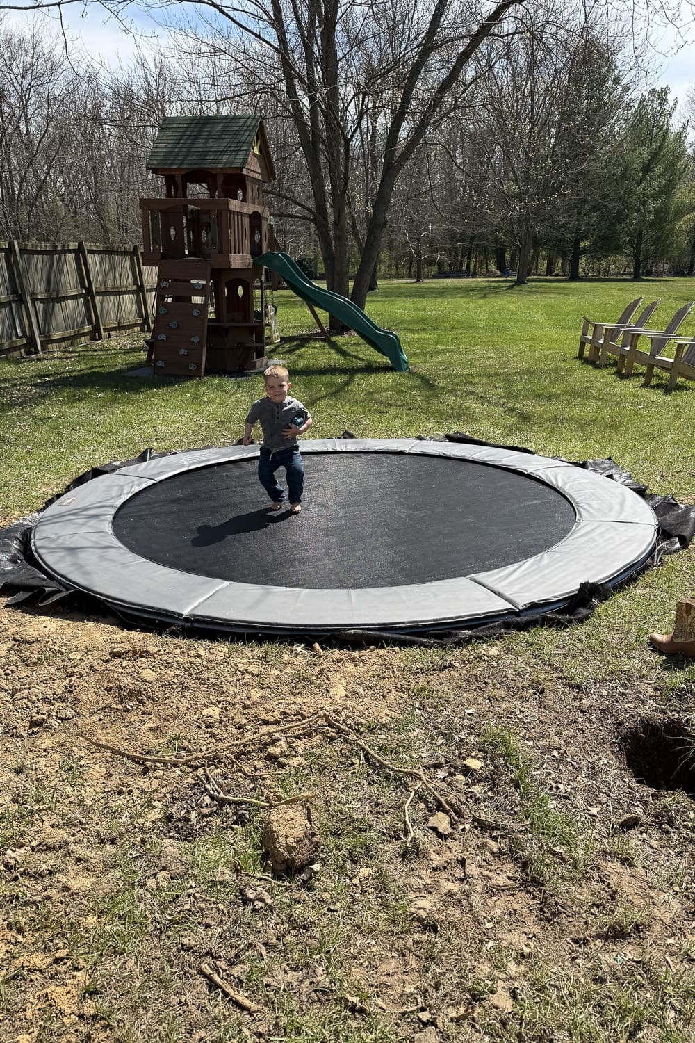 Child standing on a newly installed in-ground trampoline in a backyard with a wooden playset and open grassy yard
