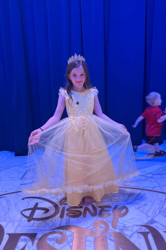 young girl wearing yellow princess dress and tiara posing on disney cruise ship during themed night with disney logo floor