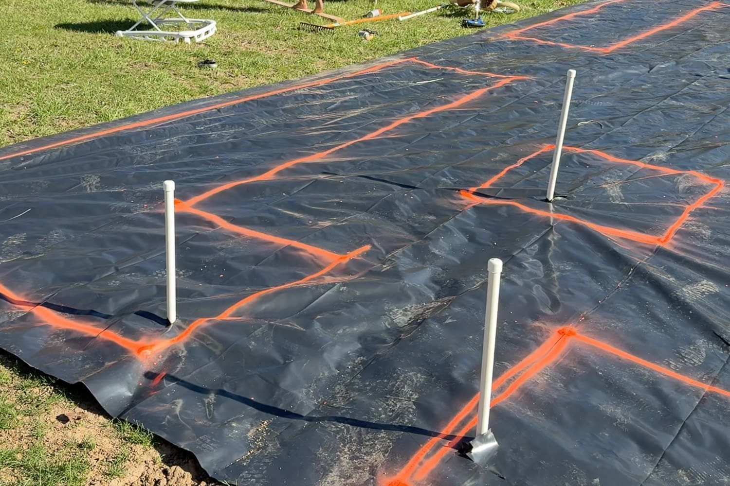 Landscape fabric laid over the buried PVC watering system with PVC spigot risers poking through and orange spray paint marking out the raised garden bed layout