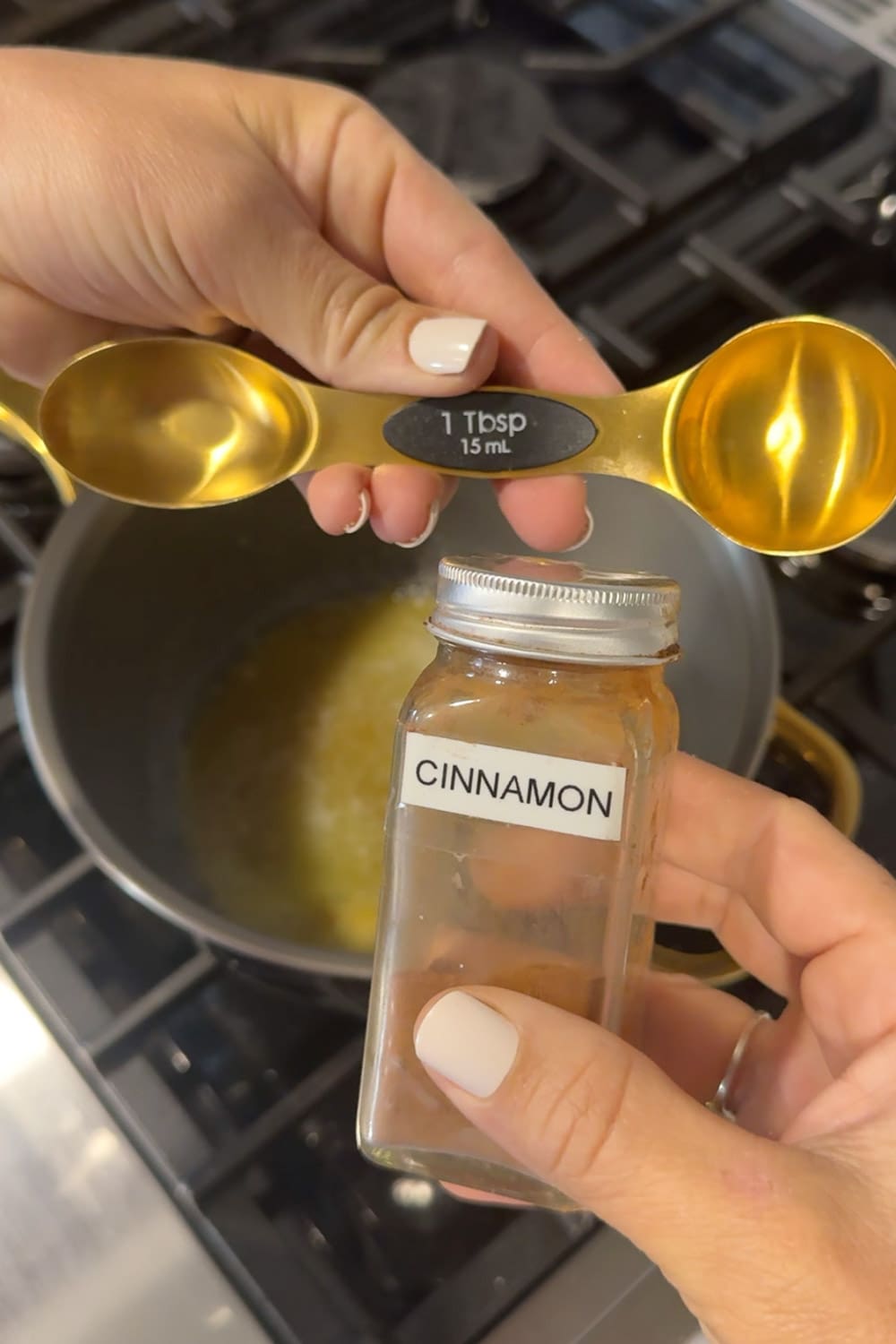 Hands holding a gold double-sided measuring spoon and a glass cinnamon spice jar over a saucepan of melted butter and brown sugar on a gas stovetop, measuring out 1 tablespoon of cinnamon for the pancake swirl filling.