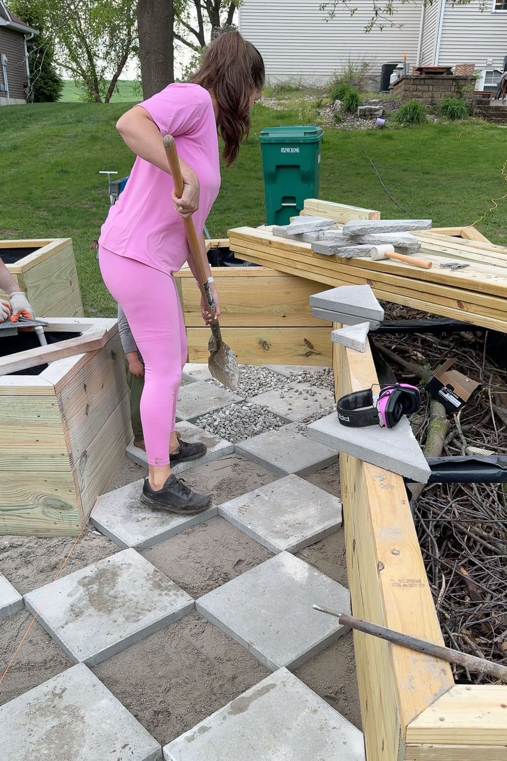 Shoveling gravel into the gaps between square pavers to complete a DIY paver walkway with a diamond checkerboard pattern
