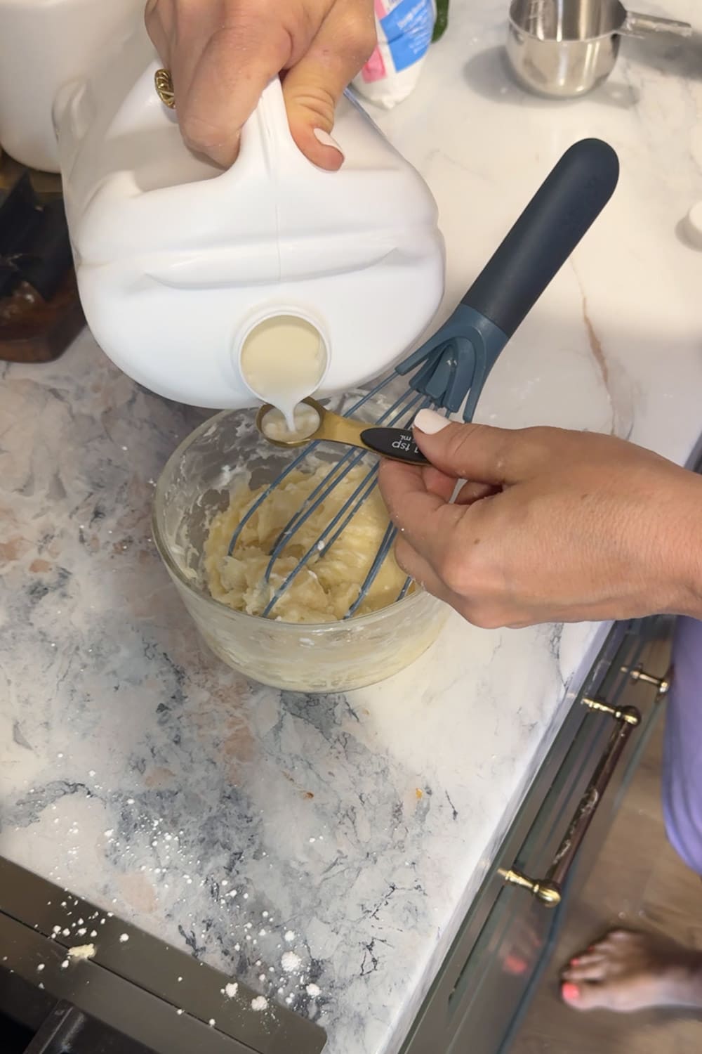 Hands pouring milk from a measuring spoon into a glass bowl of cream cheese glaze mixture while holding a blue silicone whisk, thinning the icing.