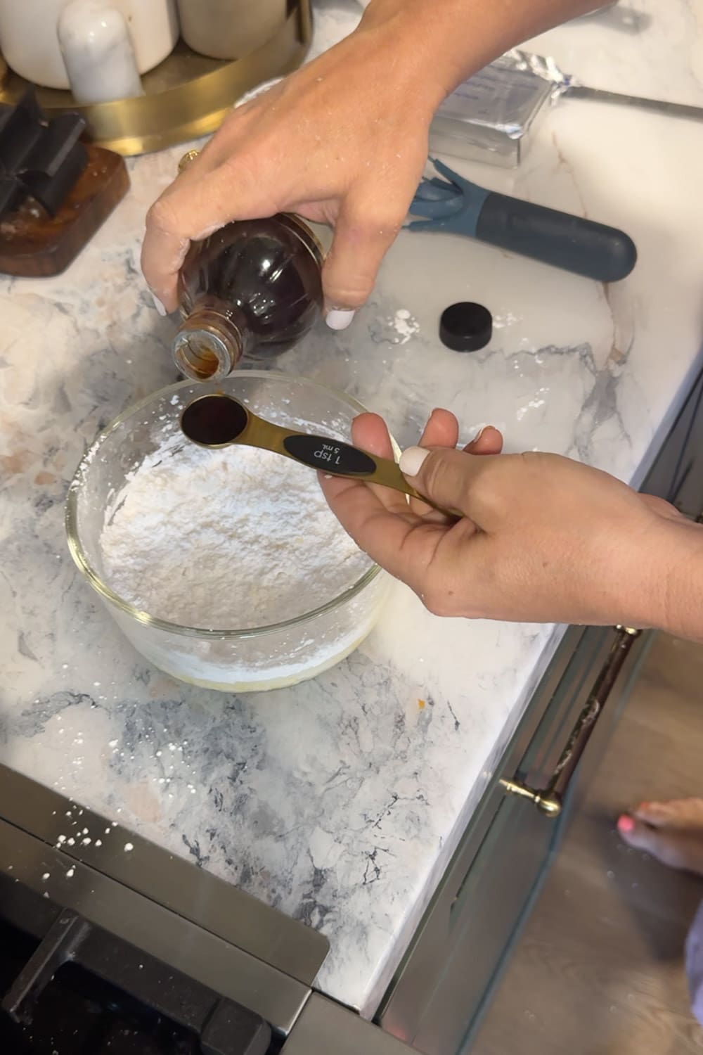 Hands measuring 1 teaspoon of vanilla extract with a gold measuring spoon over a glass bowl of powdered sugar and cream cheese, making glaze on a marble countertop.