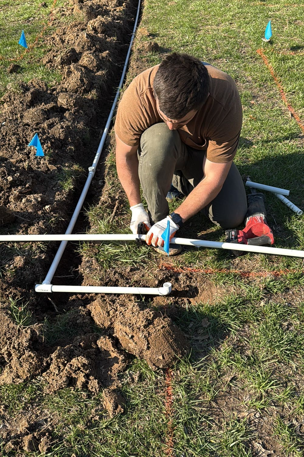 Assembling PVC pipe with a tee fitting for a DIY garden watering system, kneeling beside the trench with utility flags marking the layout