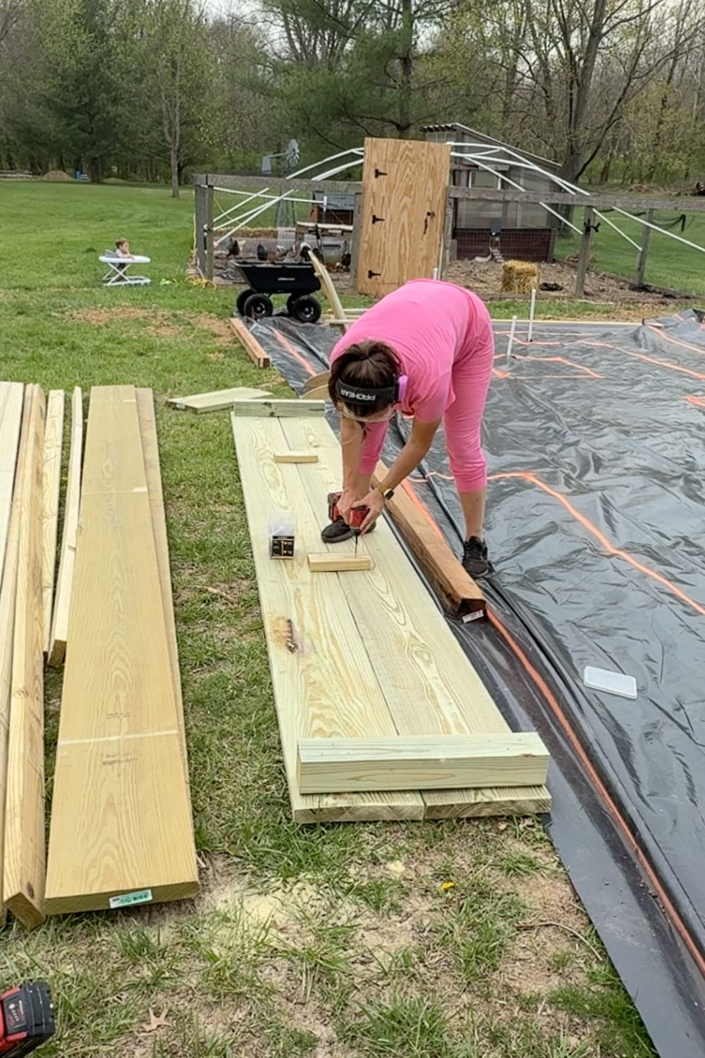 A woman in a pink outfit and pink ear protection drills screws into pressure-treated boards to assemble a raised garden bed panel, next to the spray-painted black weed barrier layout, with a chicken coop and garden cart visible in the background.