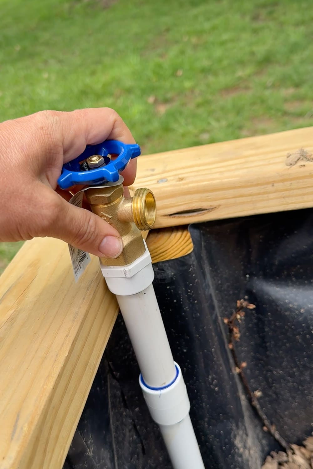 Hand attaching a brass spigot valve with a blue handle to the top of a PVC riser at the corner of a raised garden bed