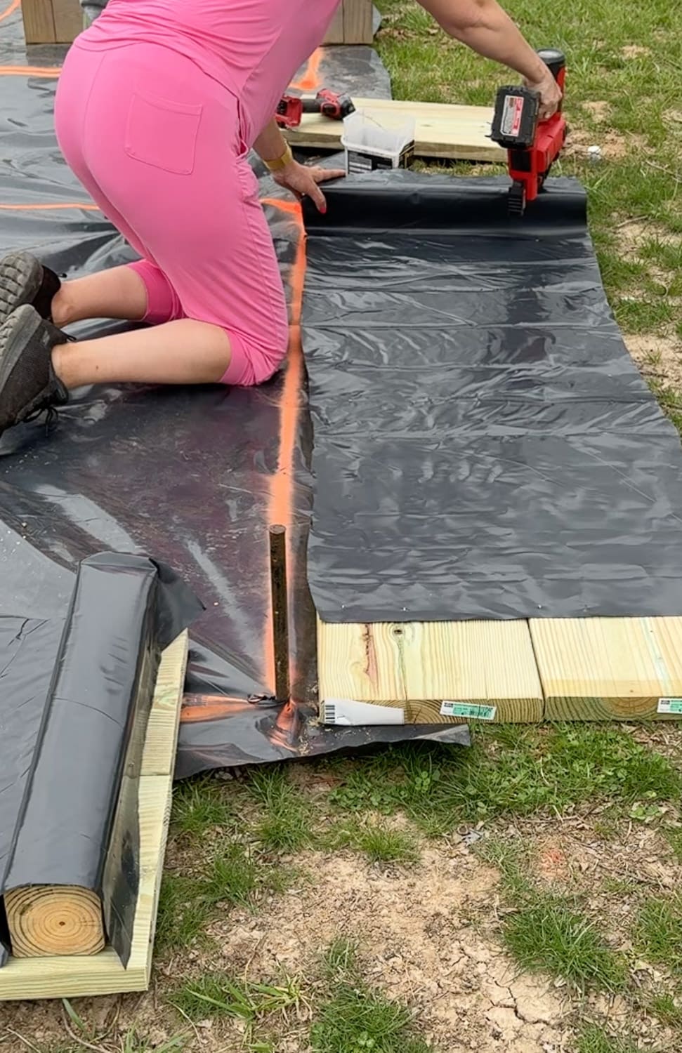 A woman in a pink outfit kneels on black weed barrier fabric and uses a red nail gun to attach the liner to the inside of a pressure-treated raised garden bed frame, with orange spray paint layout lines and additional lumber visible nearby.
