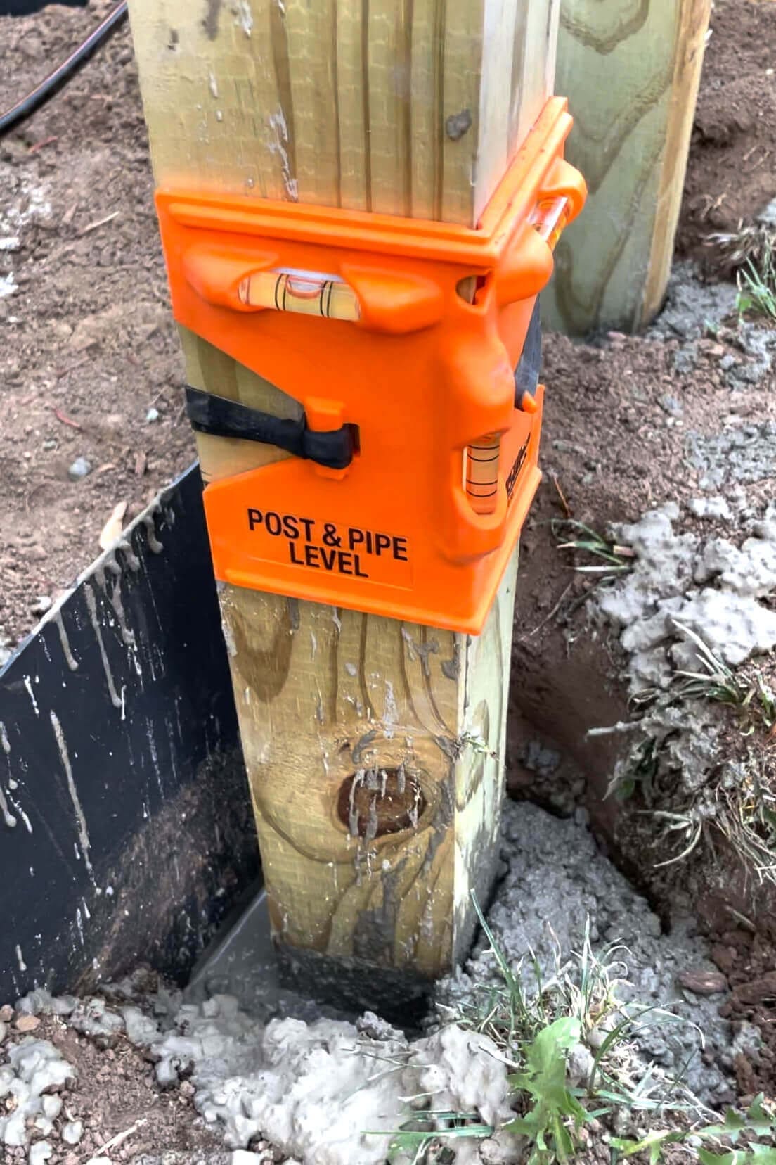 A wooden post secured in wet concrete with a bright orange post and pipe level attached to ensure it is perfectly straight. The level has built-in bubble vials for accuracy and is strapped tightly around the post. The surrounding area shows dirt, grass, and a black plastic edging barrier.