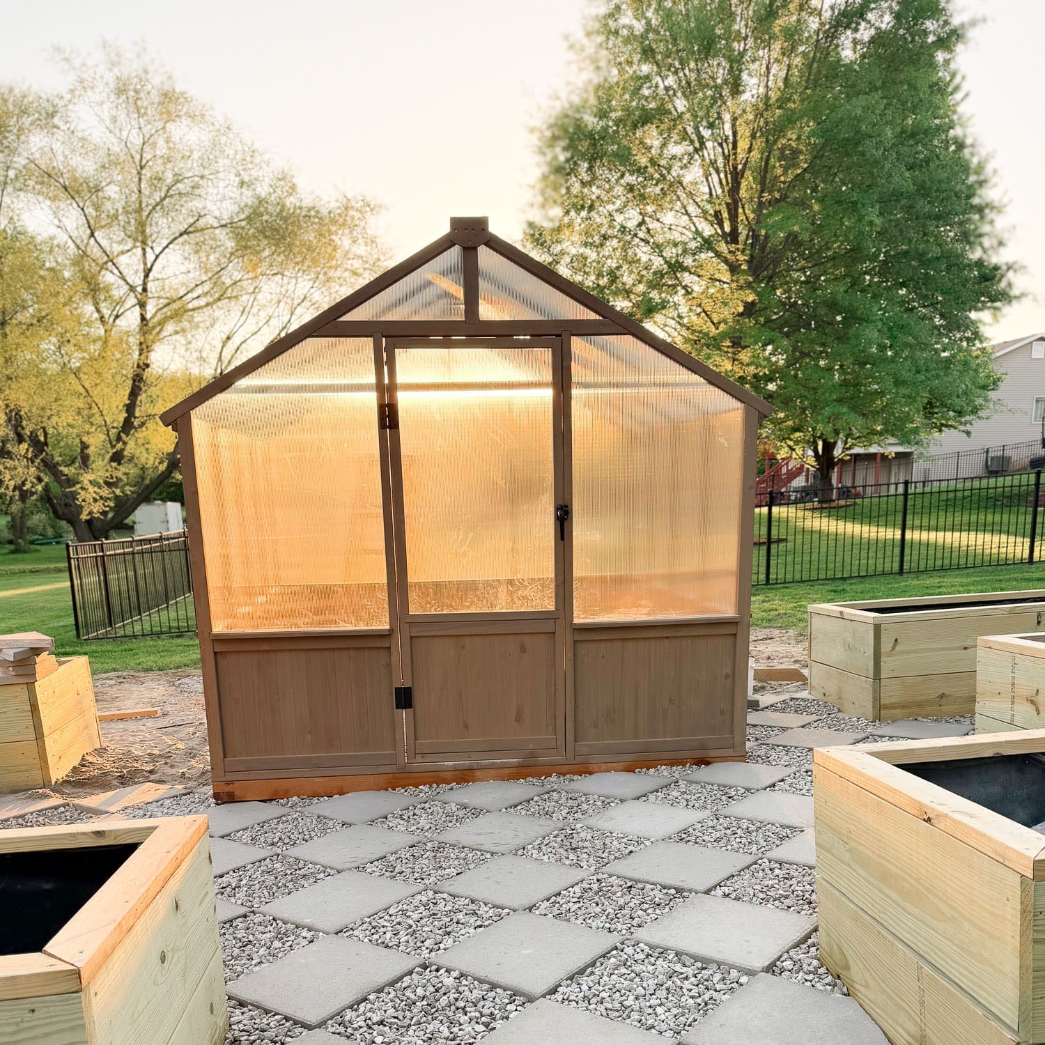 Backyard greenhouse with polycarbonate panels and wood frame, set on a checkerboard foundation of concrete pavers and gravel, surrounded by raised garden beds at sunset.