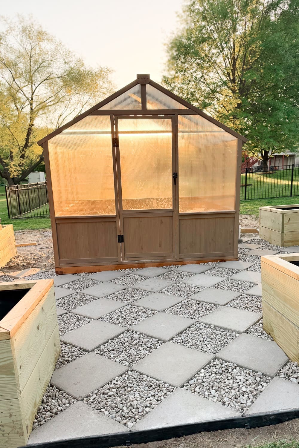 Front view of an assembled backyard greenhouse on a DIY checkerboard patio made of square concrete pavers and white gravel, with wooden raised beds on either side.