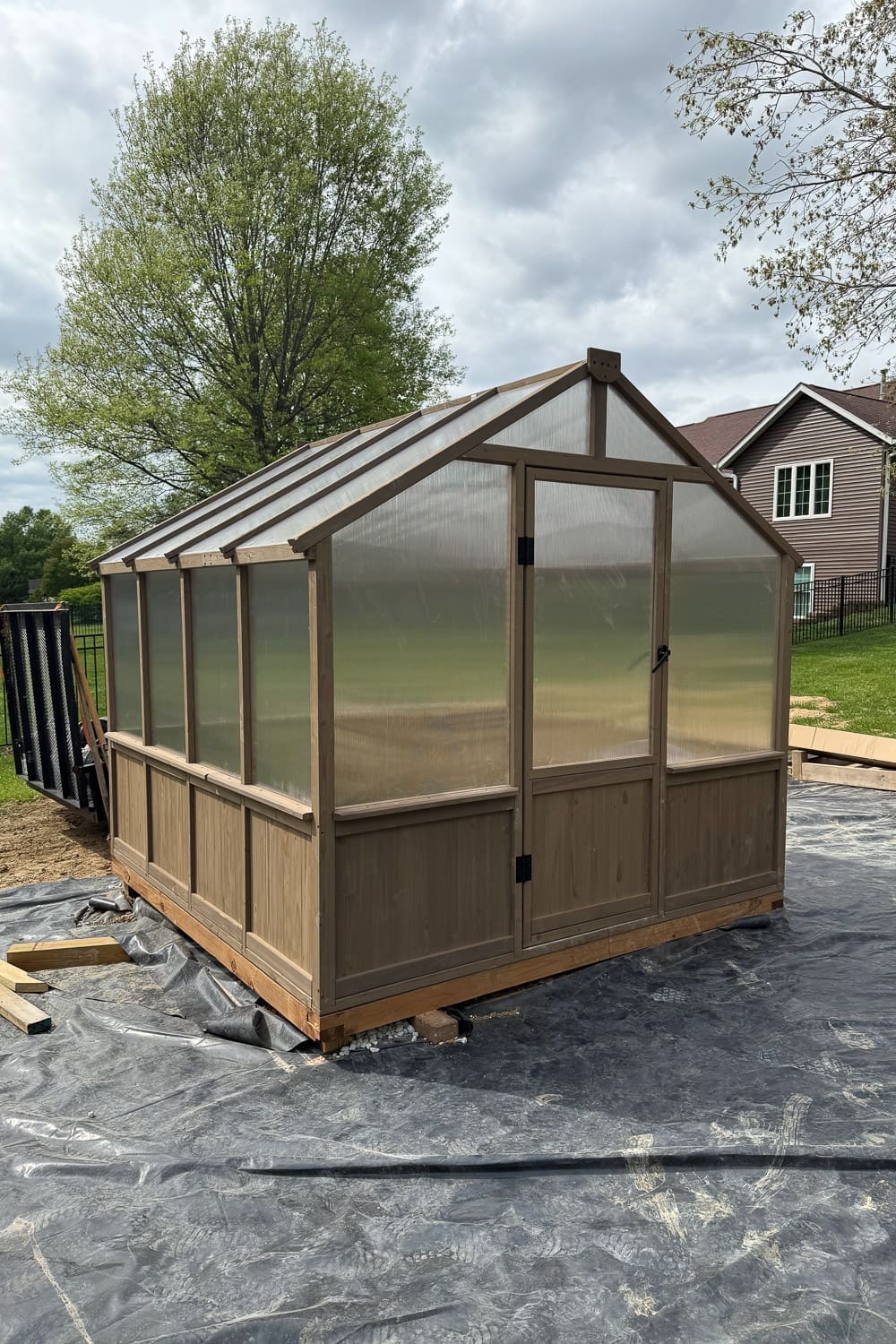 Completed backyard greenhouse with wood frame and polycarbonate panels, featuring double doors and wood lower paneling, sitting on landscape fabric before the final patio install.