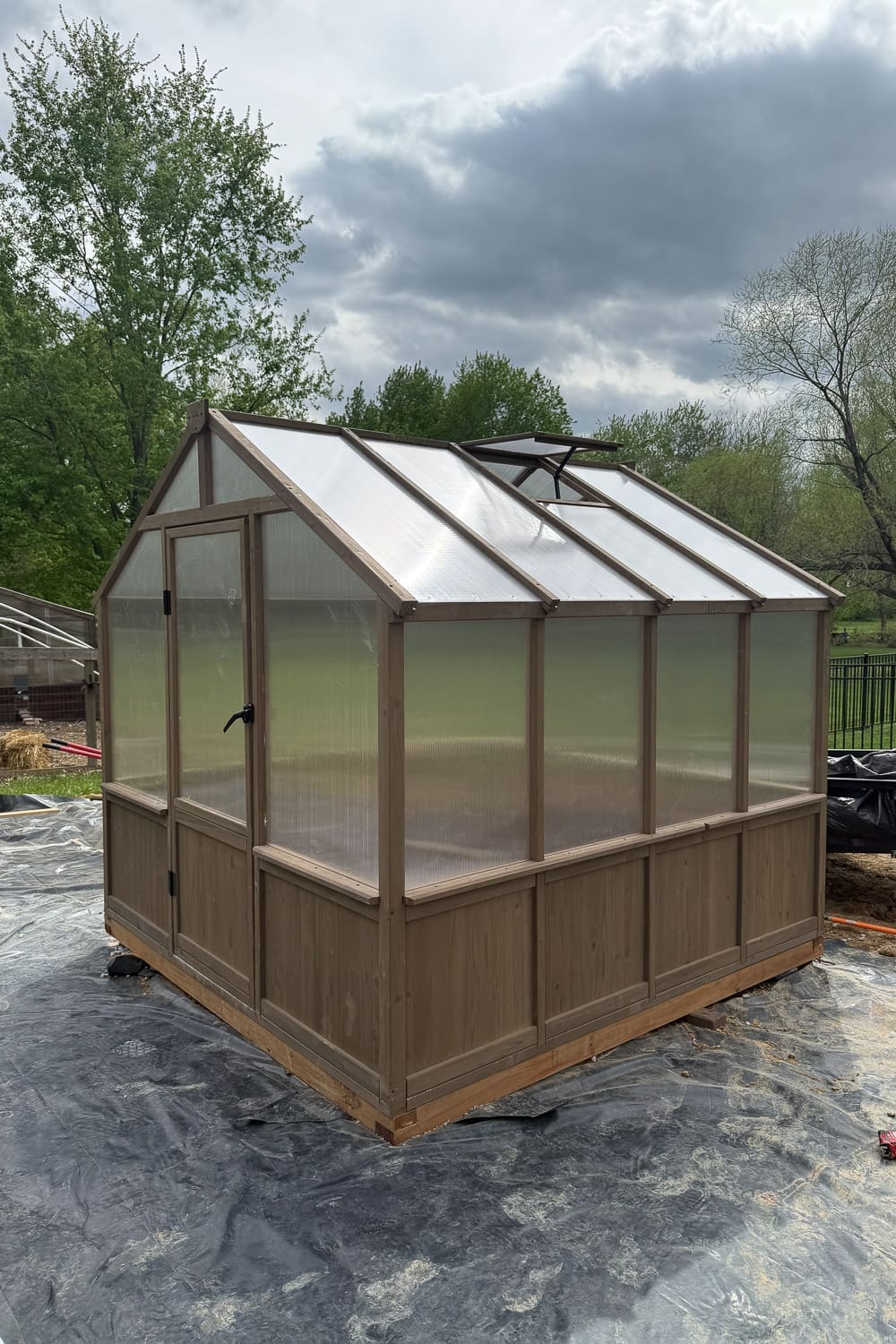 Side view of a finished backyard greenhouse showing the polycarbonate roof panels, open roof vent for ventilation, and cedar-toned wood frame.