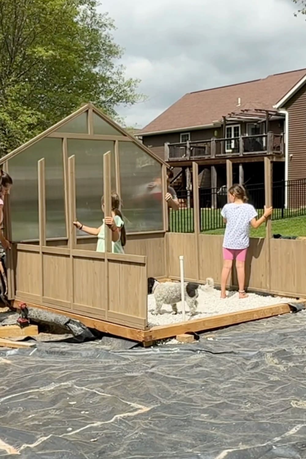 Family working together to assemble a backyard greenhouse, holding wall panels upright on the gravel foundation during installation.