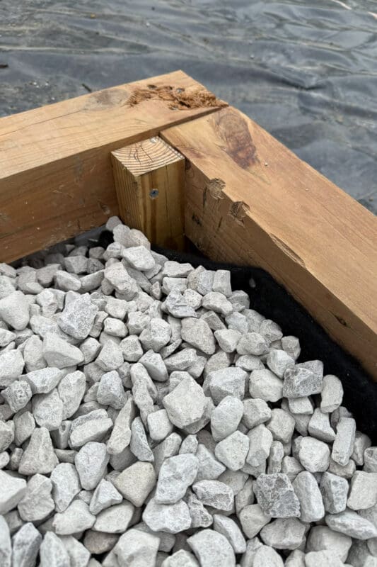 Close-up of a foundation corner showing the pressure-treated post, wood frame, landscape fabric lining, and white gravel fill.