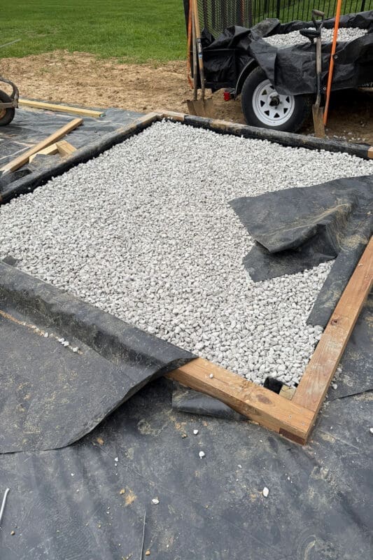 Backyard greenhouse foundation being filled with white gravel over black landscape fabric, with a wheelbarrow of stone and shovels nearby on the grass.