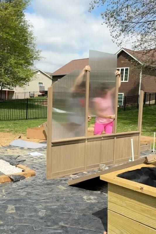Installing a polycarbonate panel into the wood frame of a backyard greenhouse during assembly, with the lower wood paneling and foundation already in place.