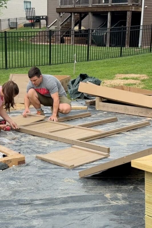 Assembling the wood wall panels of a grow house kit on the ground before lifting them into place, with boxed parts and landscape fabric nearby.