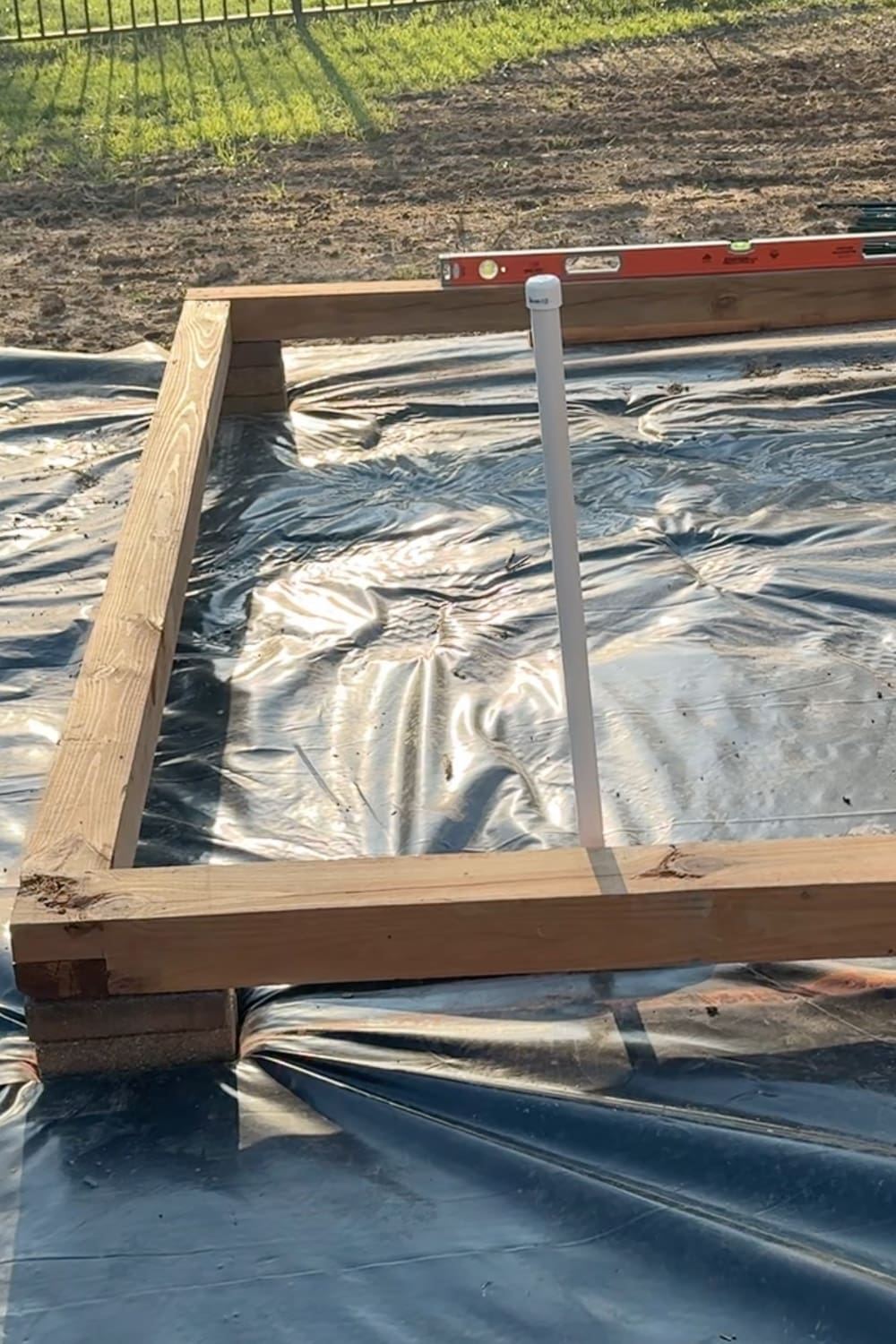 Building a wooden raised garden bed frame around a PVC watering spigot, using a level to keep the boards square on the landscape fabric base