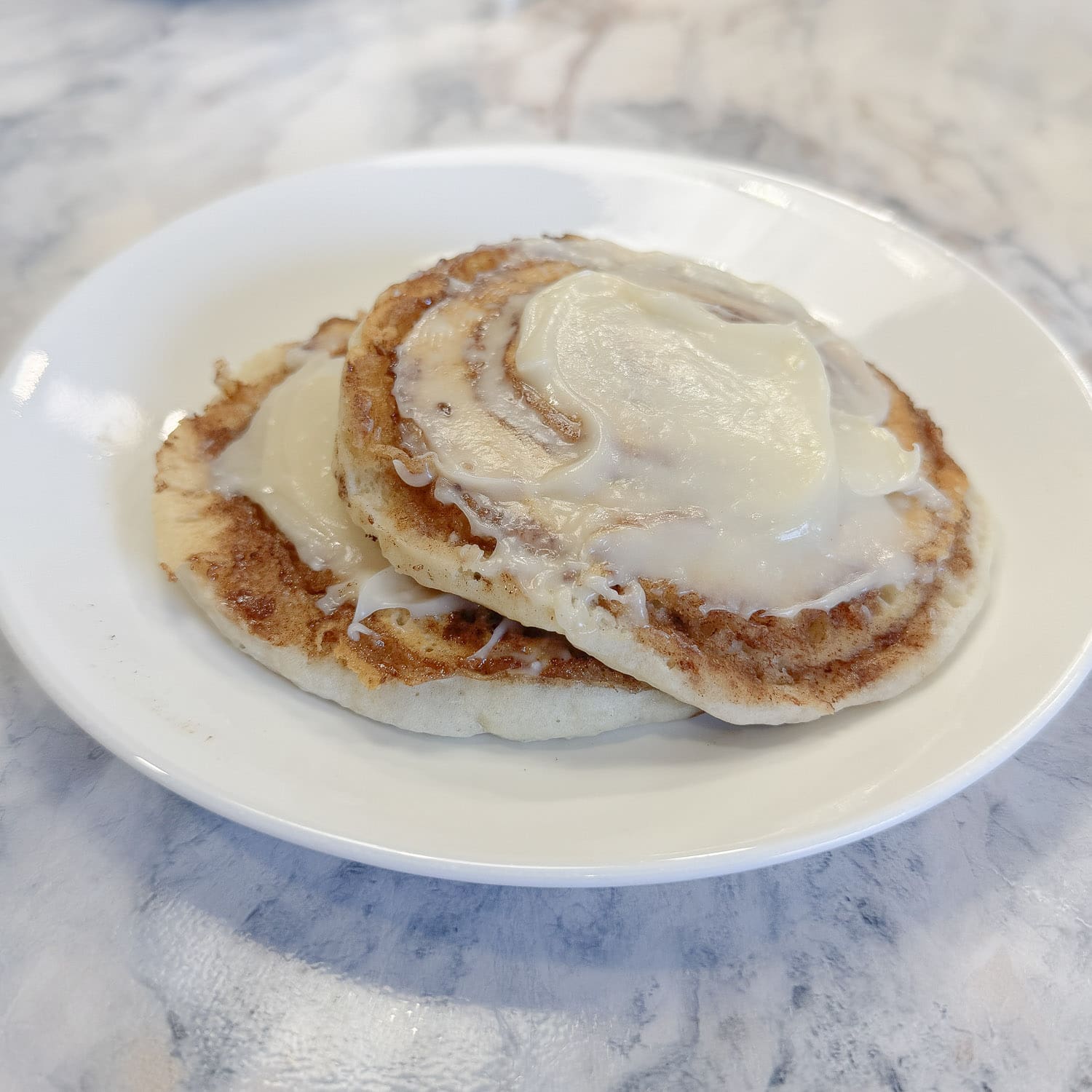 Two fluffy cinnamon roll pancakes on a white plate drizzled with cream cheese icing, with visible cinnamon swirls on the surface, photographed on a marble countertop.