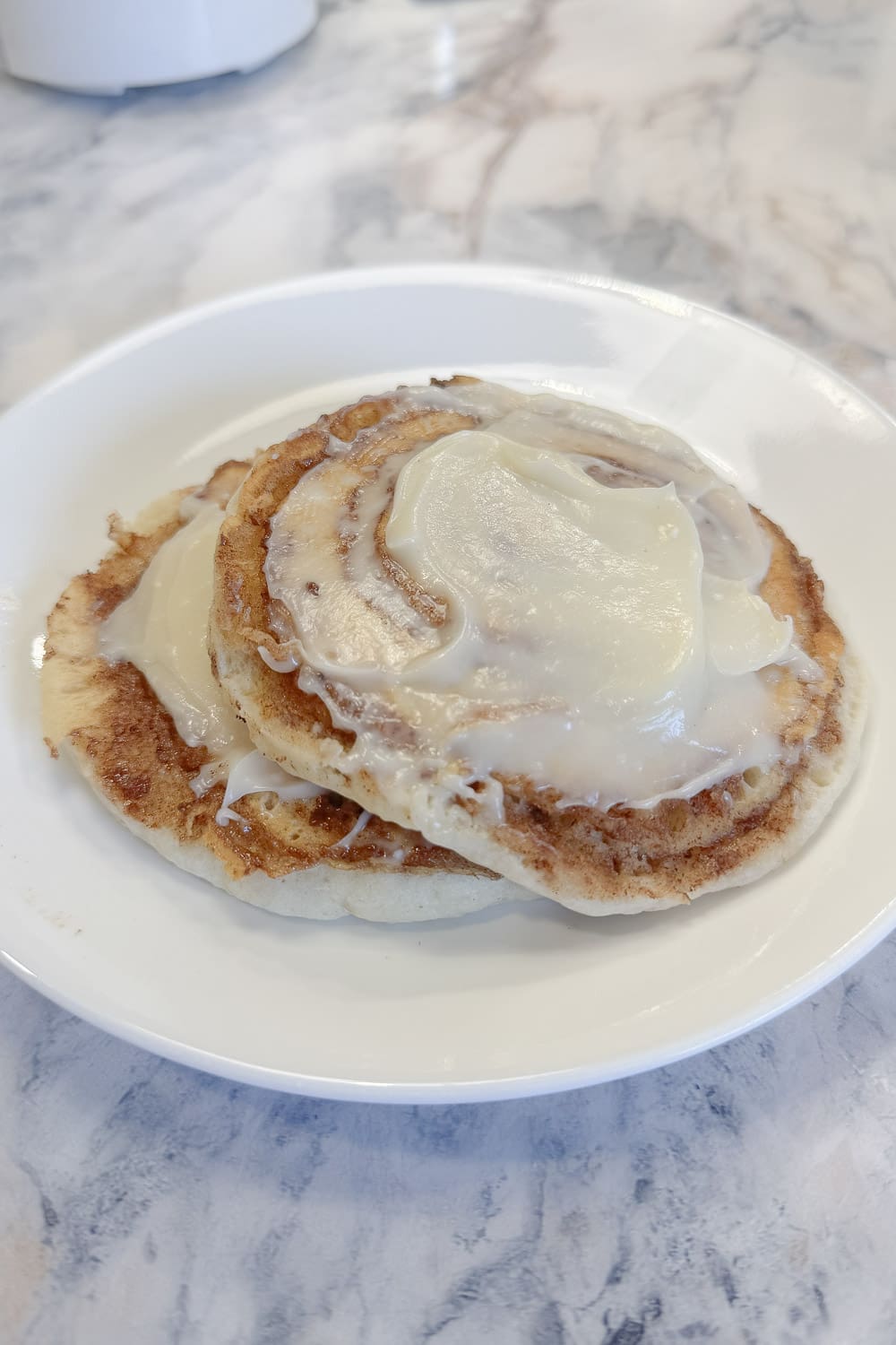 Two cinnamon roll pancakes stacked on a white plate, topped with a generous pour of melted cream cheese glaze, showing the swirled cinnamon pattern on the pancakes, on a marble countertop