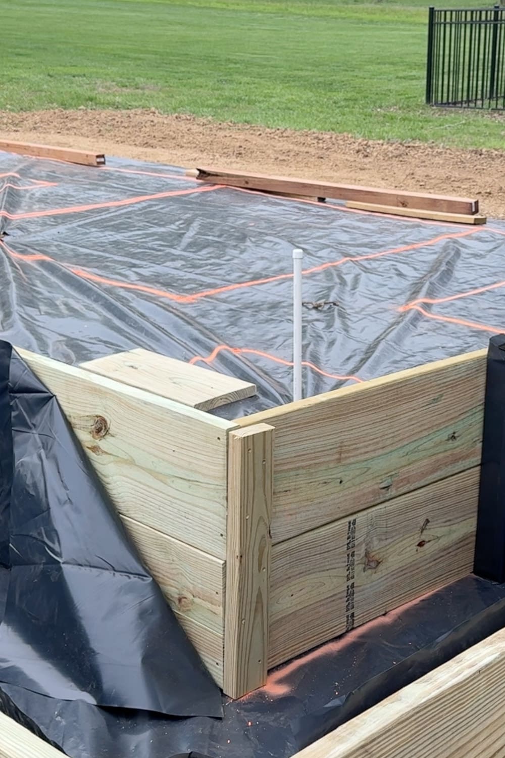 A close-up view of a completed raised garden bed corner made from stacked pressure-treated lumber, lined with black fabric, set on a weed barrier with orange spray paint layout lines. A white PVC pipe stake and additional bed outlines are visible in the background.