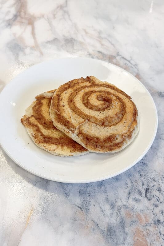 Two fully cooked cinnamon roll pancakes stacked on a white plate showing beautiful golden brown cinnamon spiral swirl patterns on top, before adding cream cheese glaze, on a marble countertop.