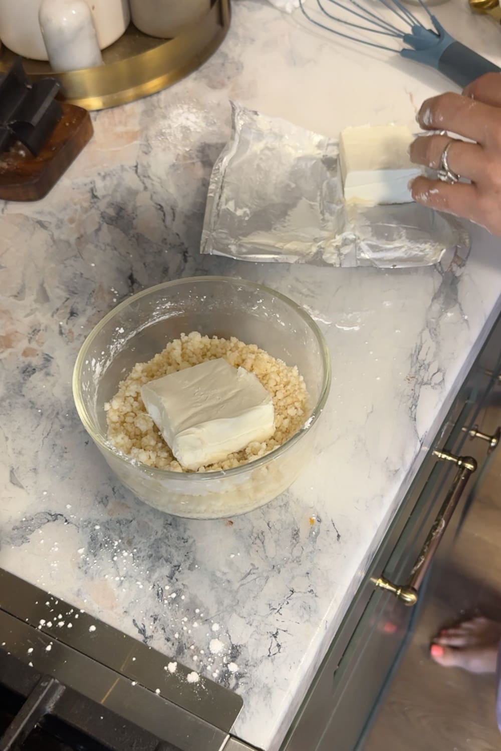 Hand placing a block of cream cheese into a glass bowl of powdered sugar on a marble countertop, showing the ingredients for homemade cream cheese glaze, with the foil wrapper set aside nearby.