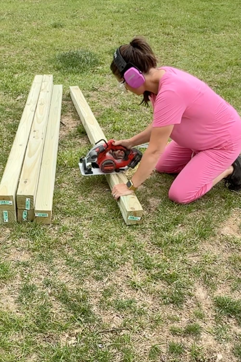 A woman in a pink outfit and pink ear protection kneels on the grass using a red circular saw to cut pressure-treated lumber, with additional uncut boards stacked nearby.