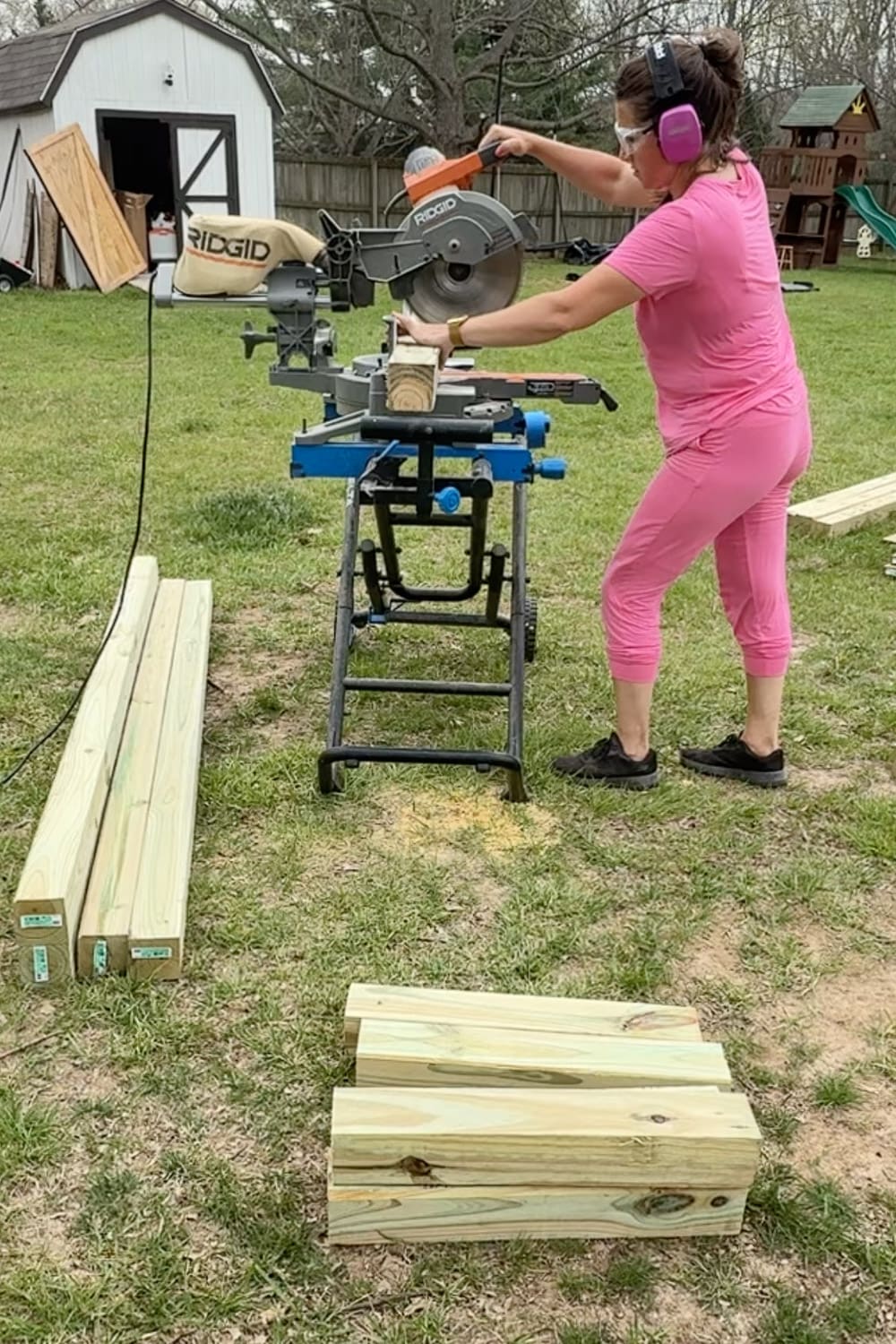 A woman in a pink outfit, safety glasses, and pink ear protection uses a RIDGID miter saw on a stand to cut pressure-treated lumber outdoors, with stacked boards and freshly cut pieces on the grass nearby and a storage shed visible in the background.