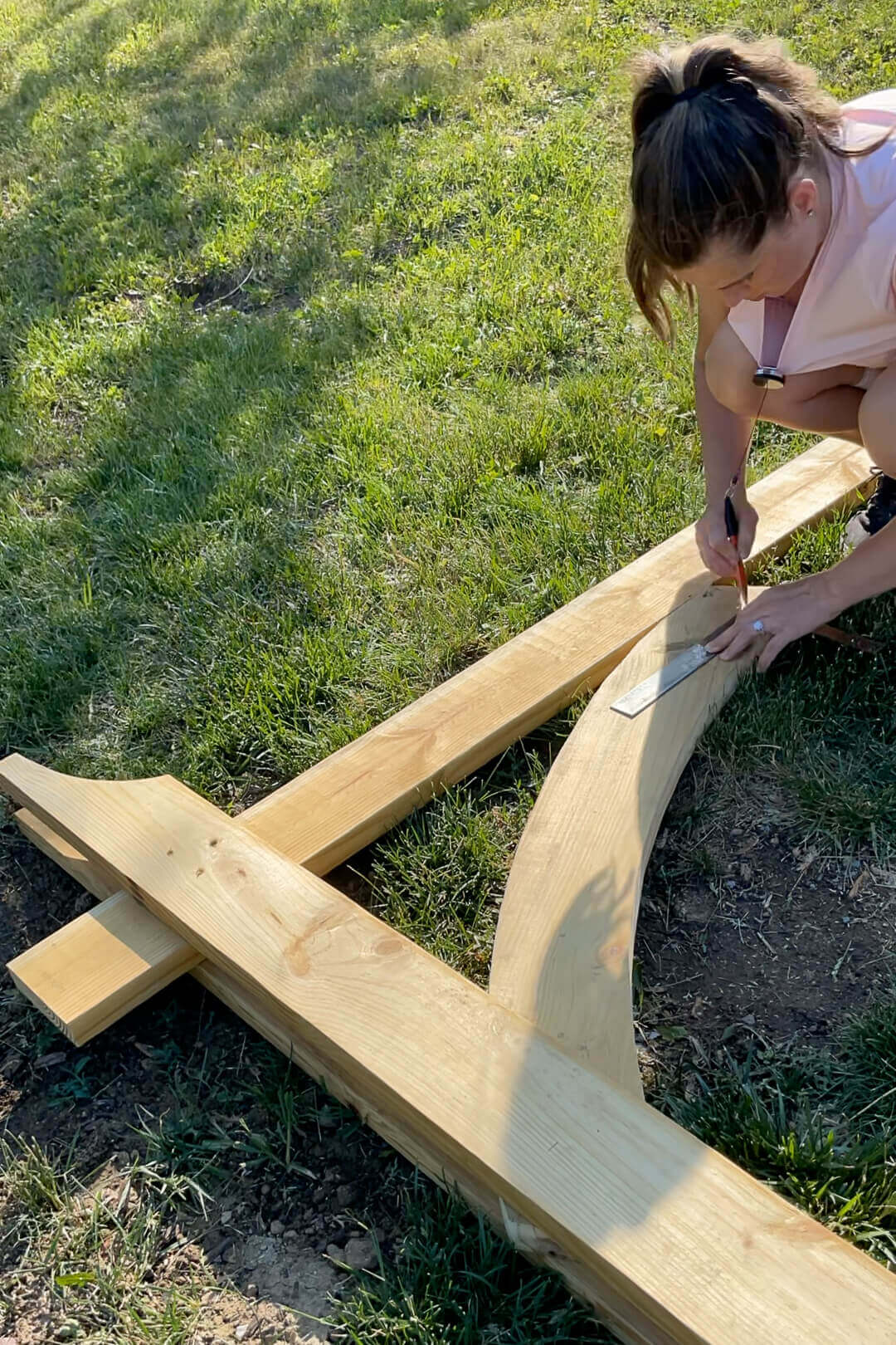 A woman in a pink shirt and black sneakers kneels on the grass, marking a wooden beam with a pencil and ruler for a precise cut. The curved design takes shape as part of an outdoor wood garden arbor.