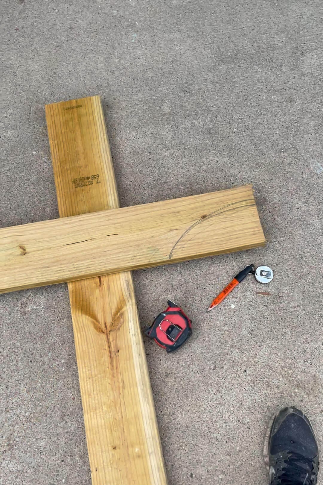 Two pieces of pressure-treated lumber crossed on a concrete surface, with a curved cut line drawn in pencil. A red tape measure, an orange carpenter’s pencil, and a metal measuring tape lay nearby, indicating a woodworking project in progress.