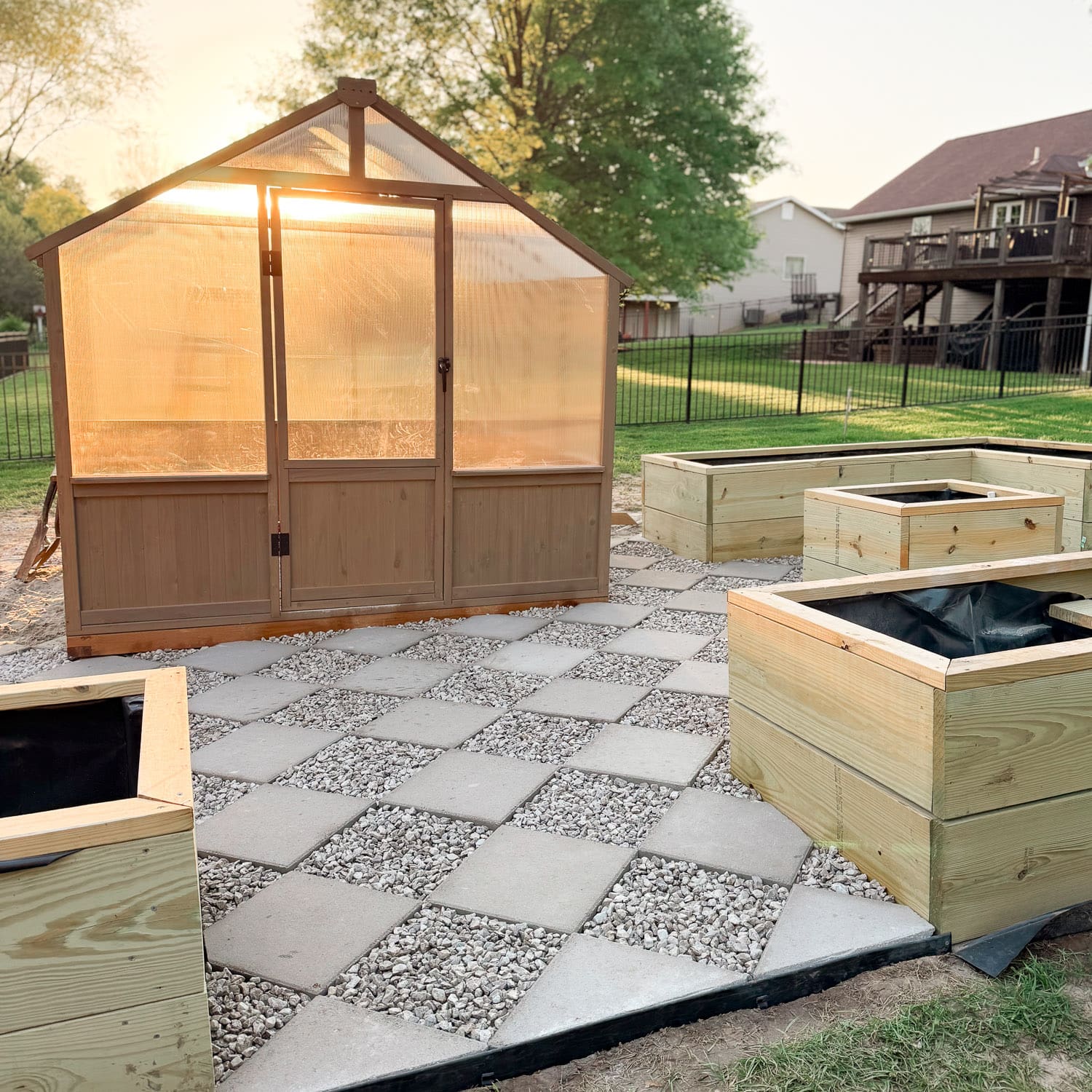 DIY paver walkway with a diamond pattern and gravel infill between raised garden boxes, with a wooden greenhouse glowing in the sunset