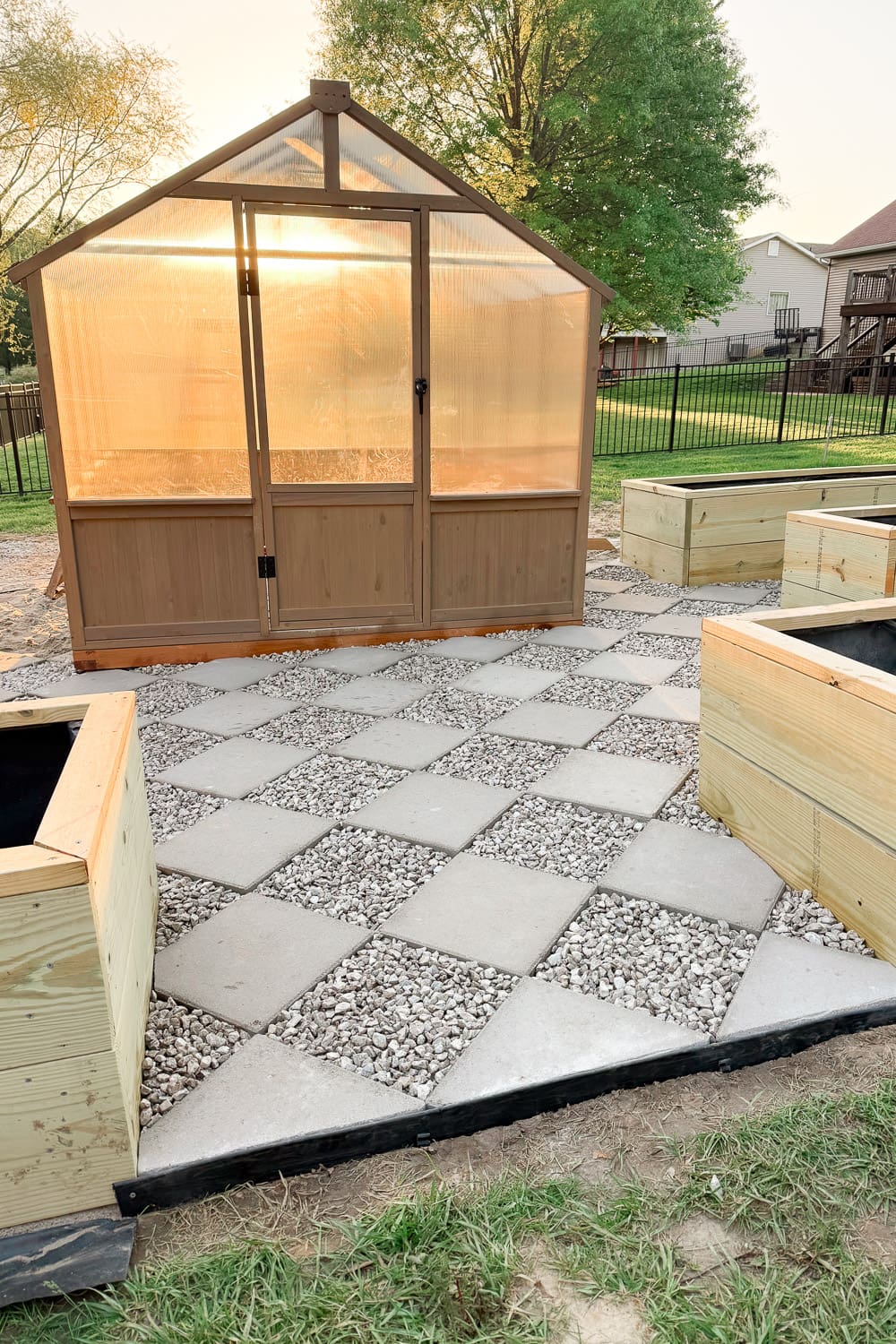 Finished DIY paver walkway in a diamond checkerboard pattern with gravel between square pavers, surrounded by raised garden beds and a greenhouse