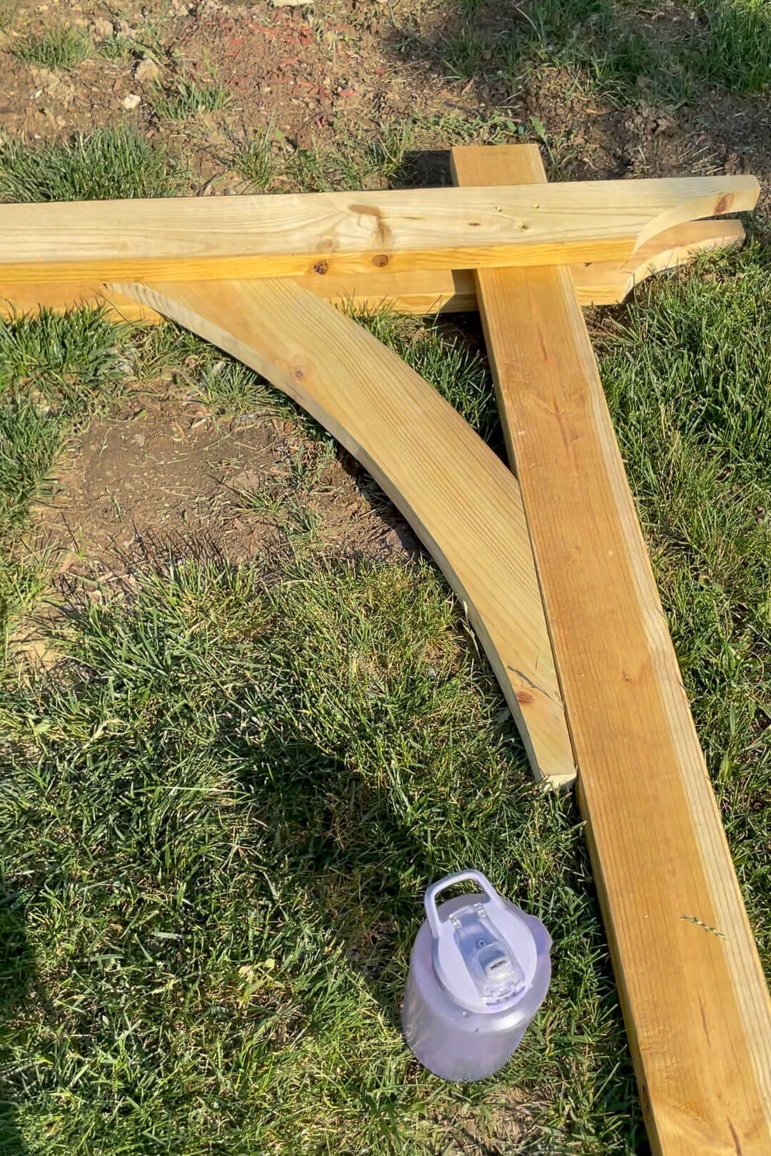 A partially assembled wooden garden arbor structure lays on the grass, with a large water bottle beside it. The curved beam is securely attached, showing progress in the DIY project.